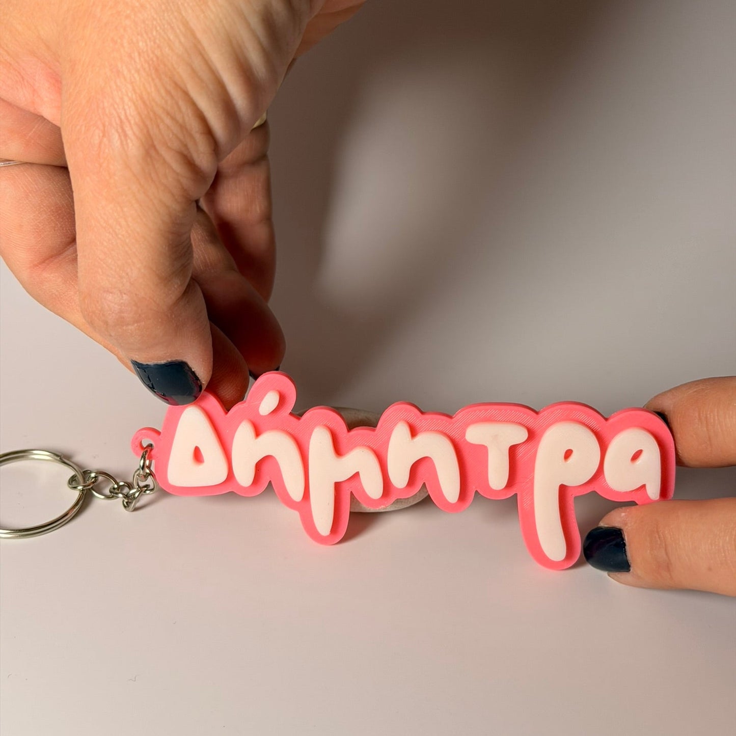 γυναικέιο χέρι κρατάει ένα custom 3D printed μπρελόκ με όνομα, φτιαγμένο από ανθεκτικό υλικό PLA με εύχρηστο κούμπωμα καραμπίνας, σε ασπρο φόντο.